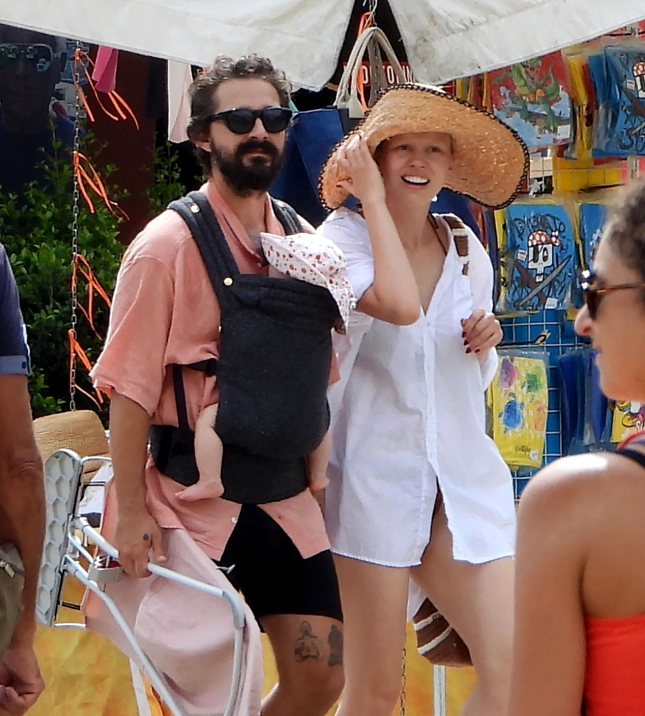 Shia LaBeouf carries his baby in a carrier next to Mia Goth, who is wearing a straw hat.