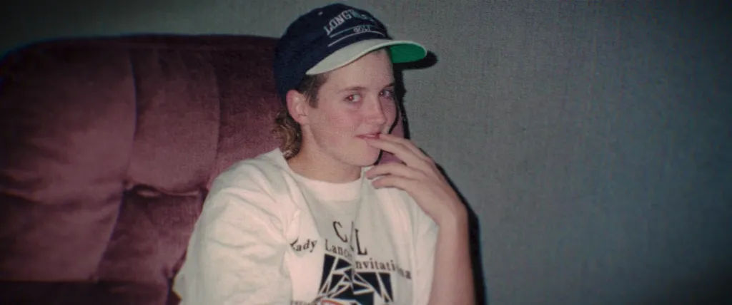 Amy Bradley, wearing a baseball cap and white t-shirt, sits on a maroon couch.