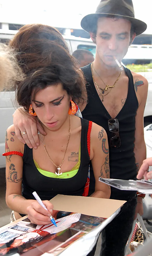 Amy Winehouse signing an autograph while a man smokes behind her.