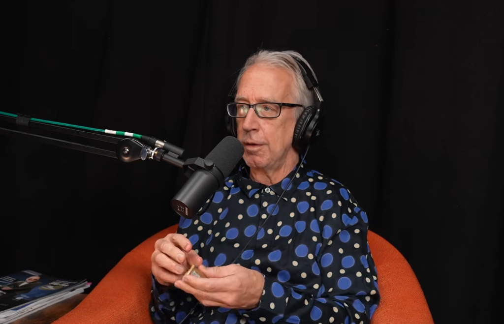 Andy Dick, wearing headphones and glasses, speaks into a microphone while holding a small object.
