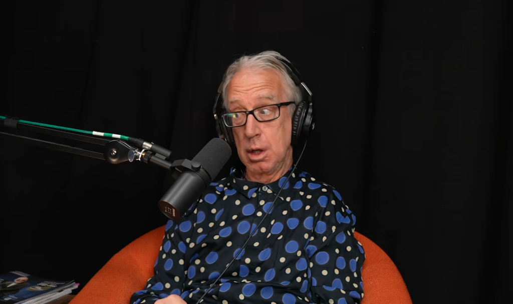 Andy Dick talking into a microphone, wearing glasses, headphones, and a blue polka-dot shirt.