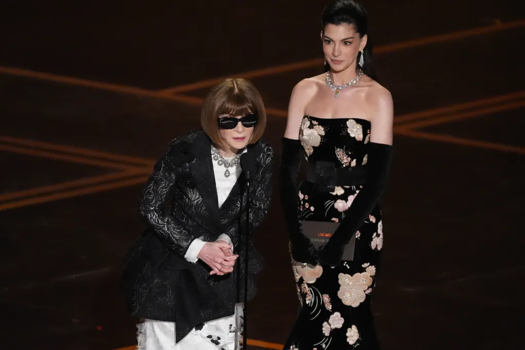 Anna Wintou, in a black lacy jacket over a white satin gown, and Anne Hathaway, in a black strapless floral gown and black gloves, present the award for best costume design at the Oscars.