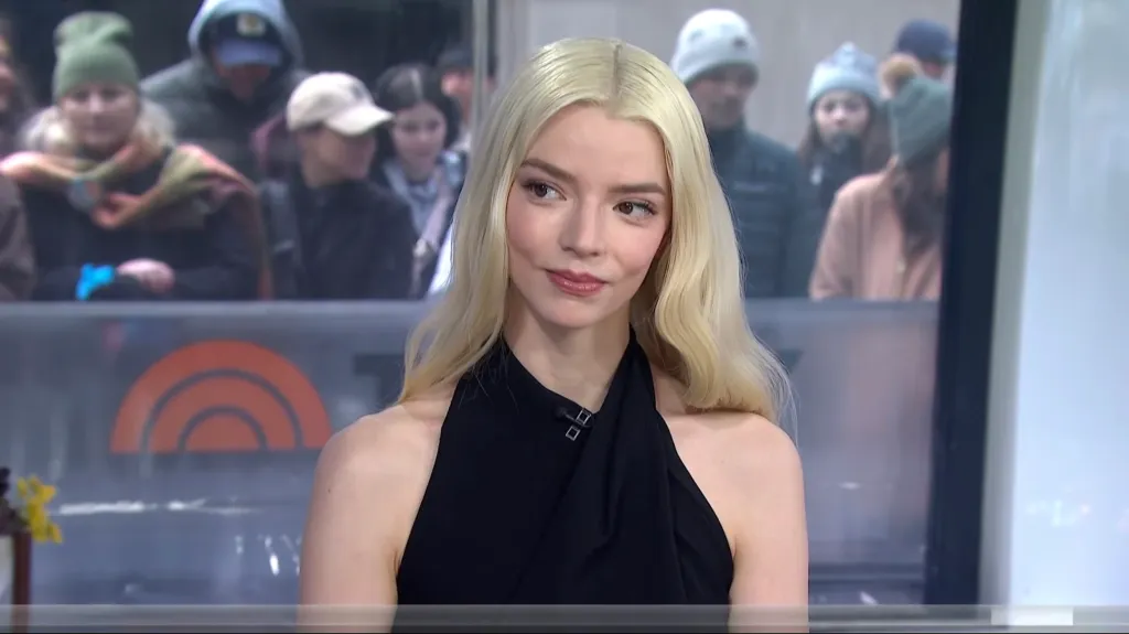 Anya Taylor-Joy in a black halter top, looking to her left with a slight smile as people stand behind a window.