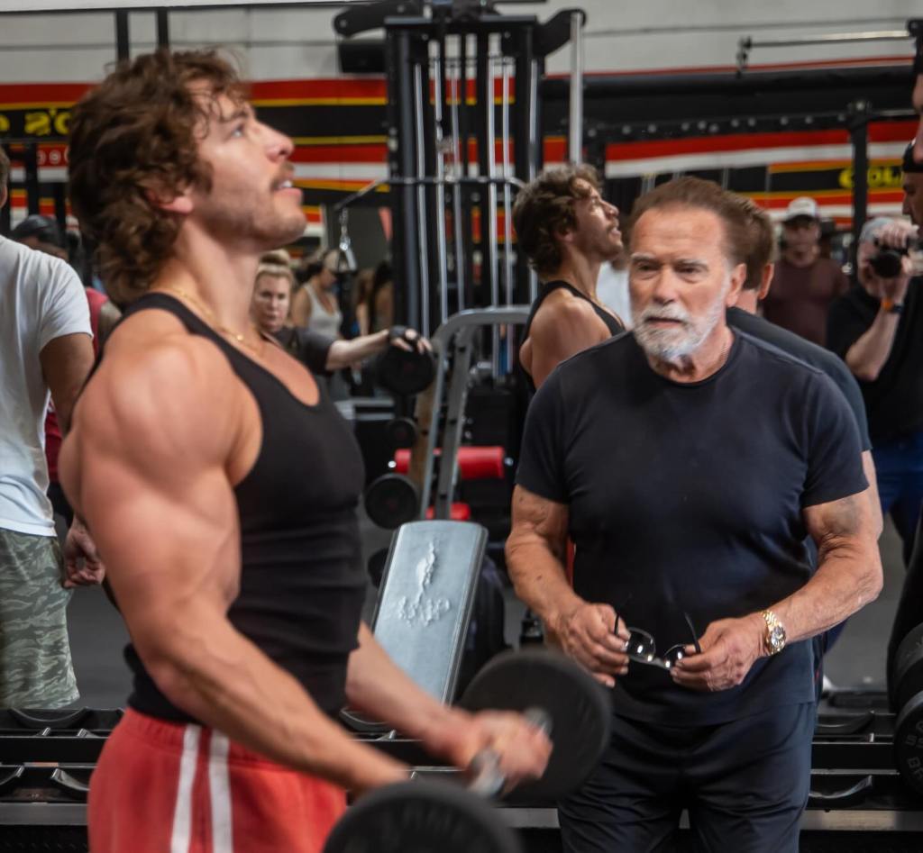 Arnold Schwarzenegger watching Joseph Baena lift weights at a gym.