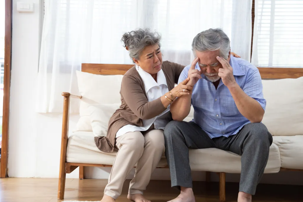 An elderly Asian woman comforts an elderly Asian man who is holding his head in distress, possibly from dementia or Alzheimer's.