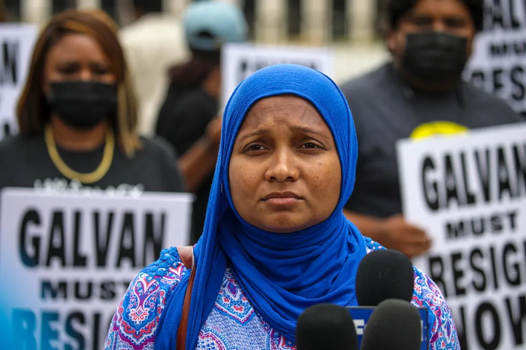 Fatima Iqbal-Zubair, wearing a blue headscarf and patterned top, speaks at a press conference with protest signs in the background.