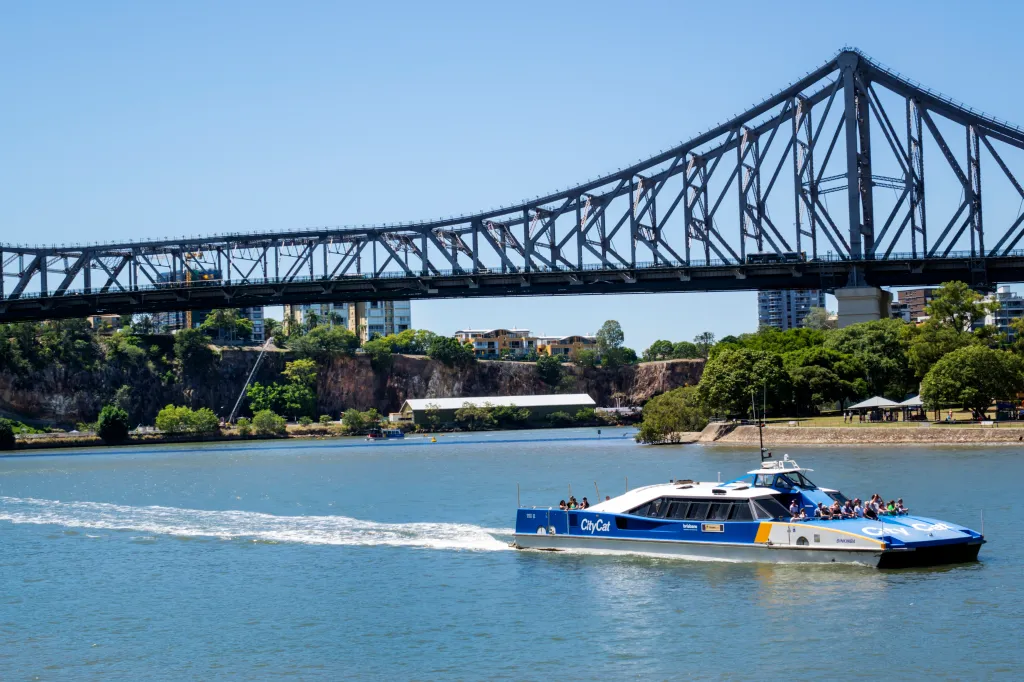 A blue and white CityCat ferry with passengers on the deck passes under the Story Bridge on the Brisbane River.