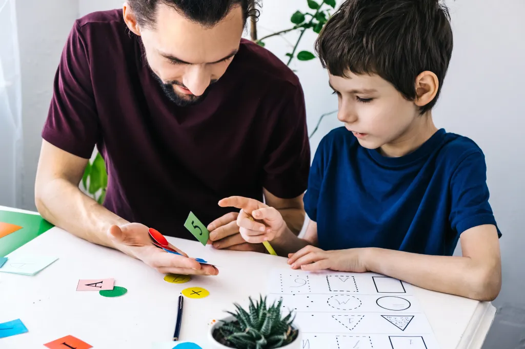 A man and a boy are learning together at a table. The man holds up a green card with the number 5, and the boy points to a worksheet with geometric shapes.