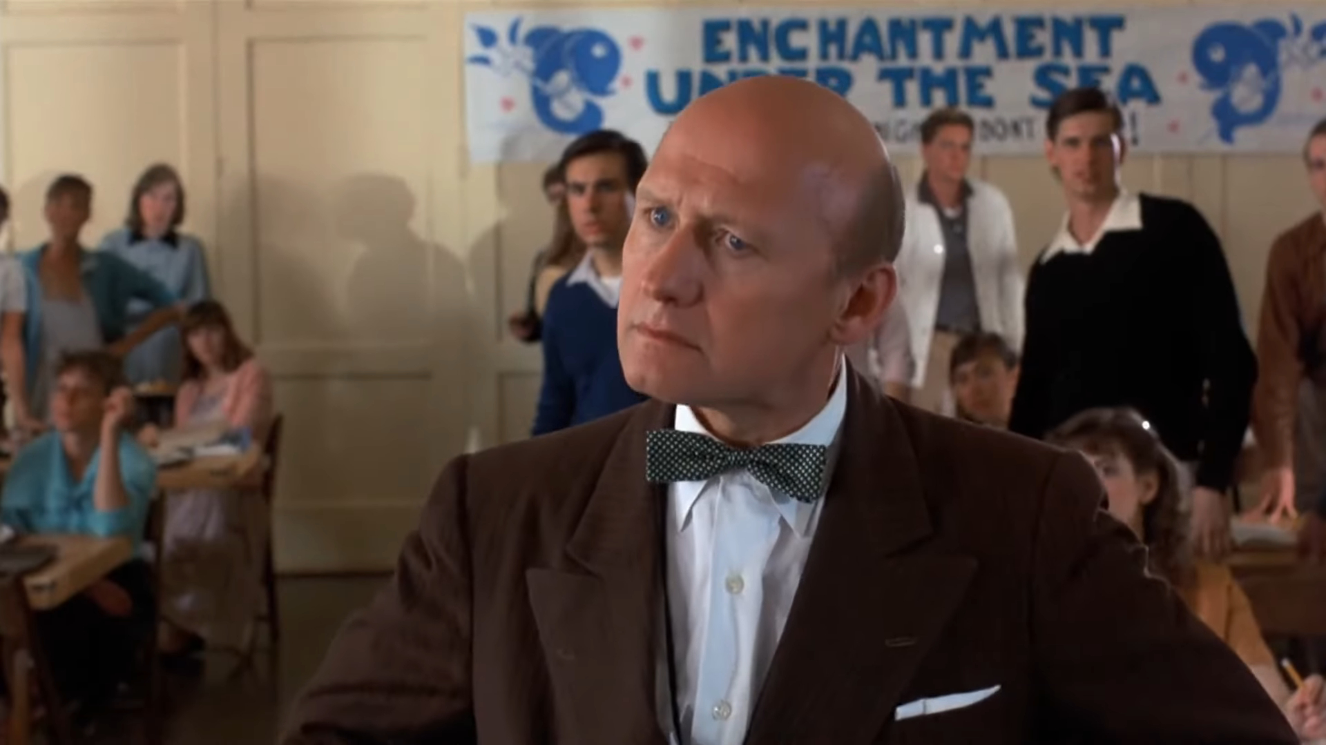 A bald man in a suit and bow tie, with students behind him and an