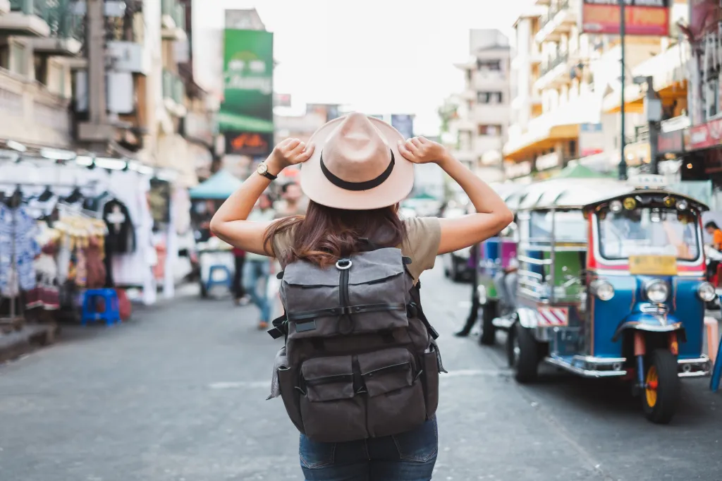 Back view of an Asian woman tourist with a backpack and hat walking on Khao San Road in Bangkok, Thailand.