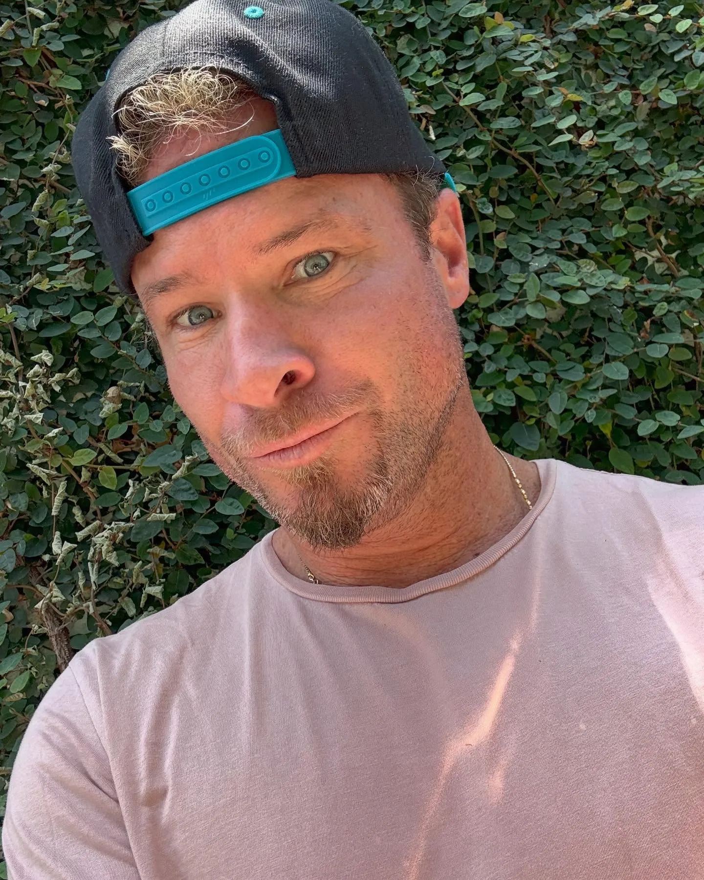 Brian Littrell from the Backstreet Boys wearing a baseball cap and pink shirt in front of an ivy wall.