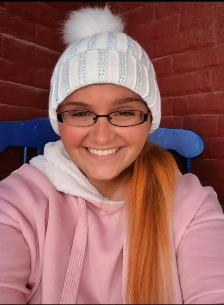 A smiling Rowan MacKenzie wearing glasses, a white knit hat with a pom-pom, and a pink hoodie.