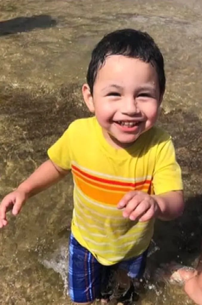 Alex Aaron Rocha, a young boy with dark hair, smiles broadly while standing in shallow water, wearing a yellow striped shirt and blue plaid shorts.