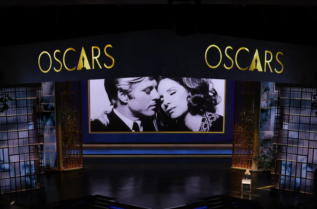 Barbra Streisand speaking at the 98th Academy Awards, with a black and white image of her and Robert Redford displayed on a large screen above her.