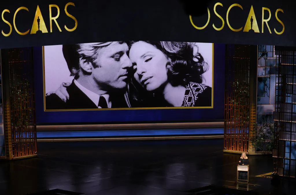 Barbra Streisand speaking at the 98th Academy Awards, with a black and white image of her and Robert Redford on a large screen behind her.