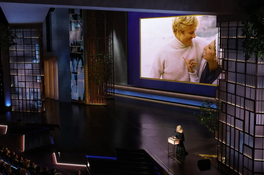 Barbra Streisand speaking onstage during the 98th Oscars, with a large screen displaying a still of her and Robert Redford from the movie *The Way We Were* in the background.