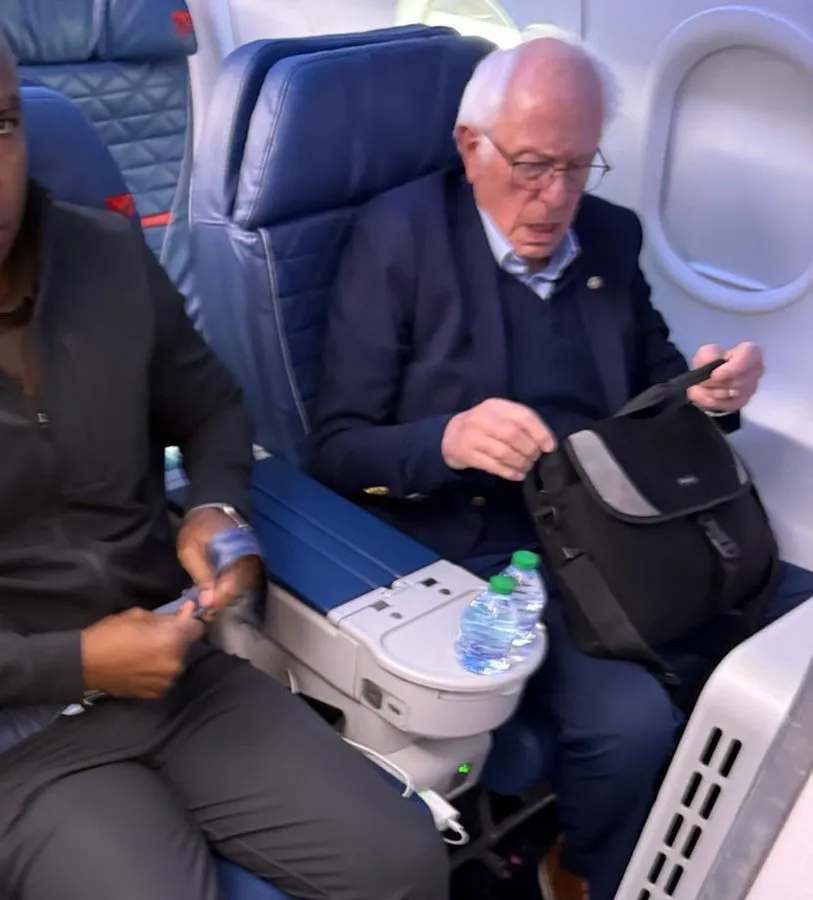 Bernie Sanders sitting in a first-class airplane seat, looking into a bag.