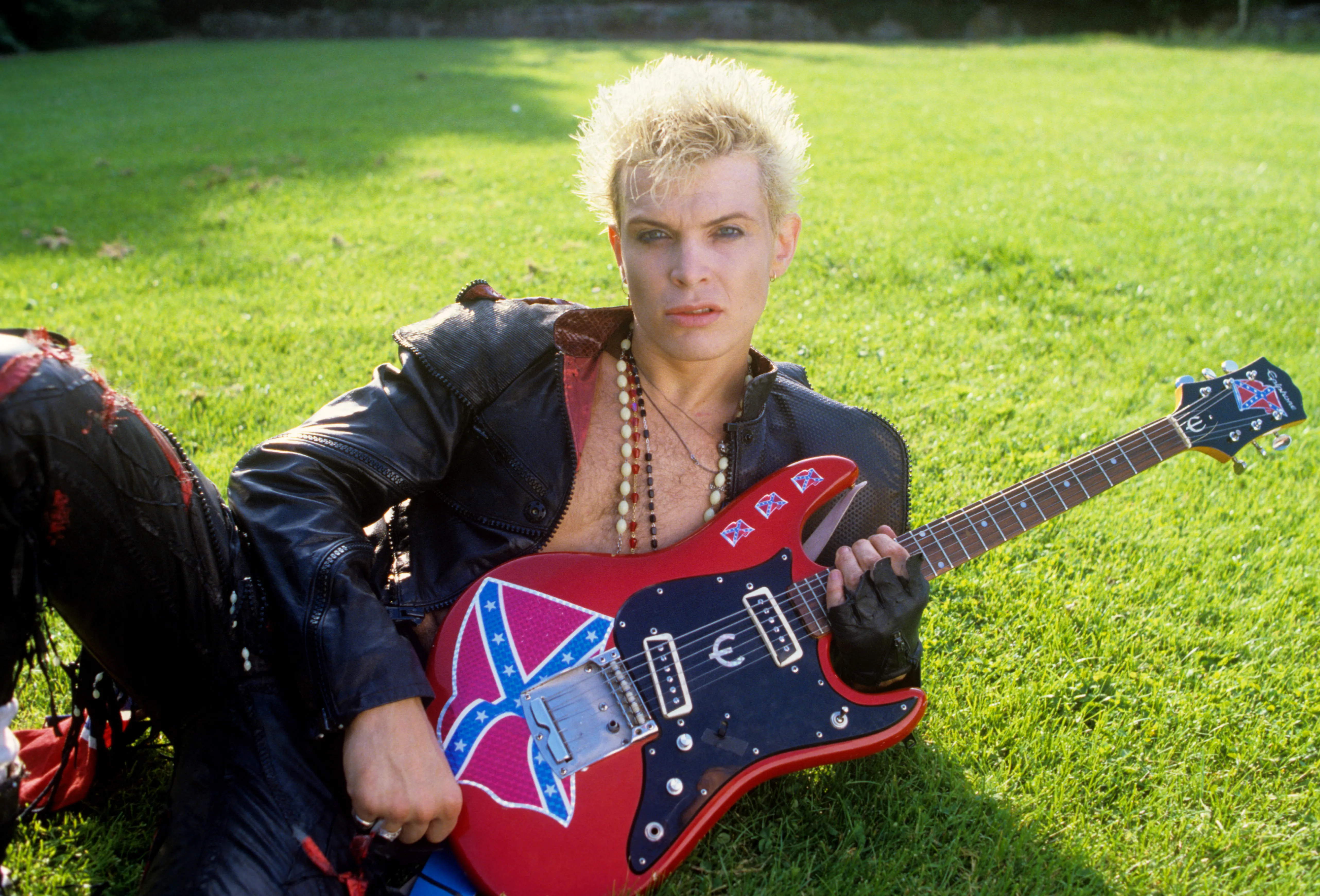 Billy Idol posing with a guitar with confederate flag sticker on it, outdoors.