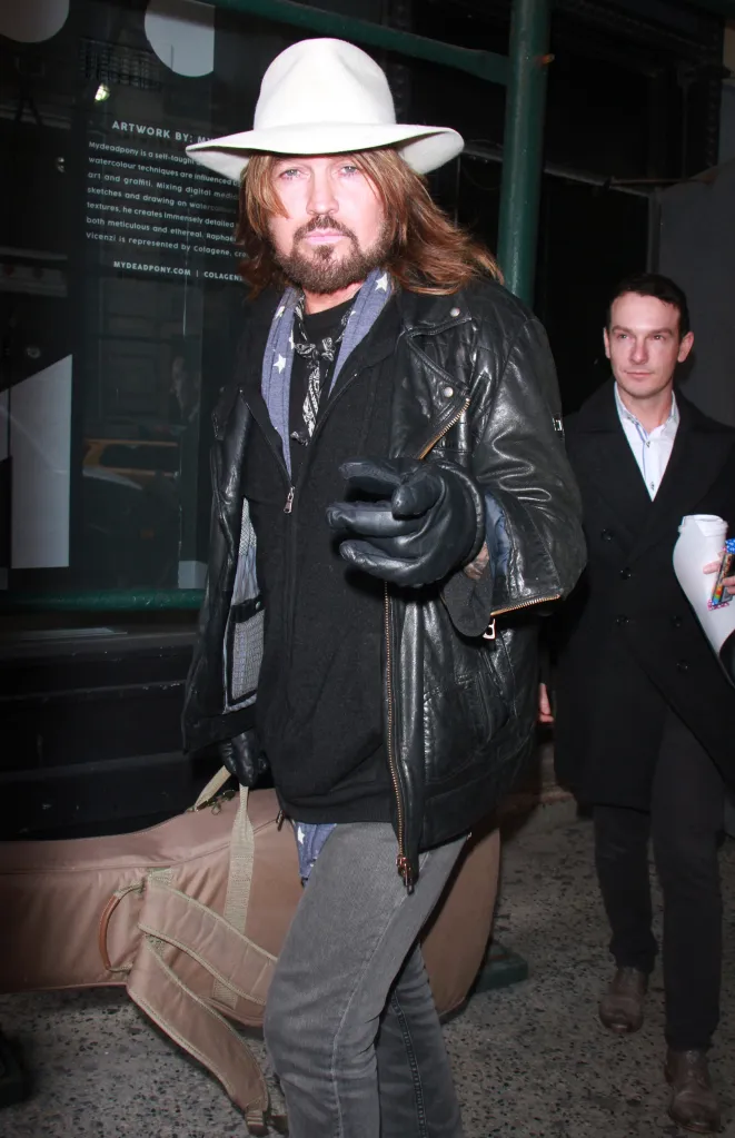 Billy Ray Cyrus wearing a white Stetson, leather jacket, and gloves, walking down a city street with a man carrying a guitar case.