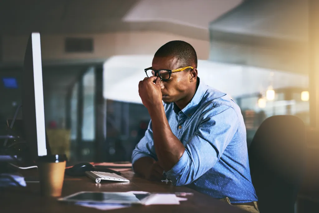 A Black man in a blue shirt and glasses sits at a desk, rubbing his temples, next to a computer and coffee cup, appearing stressed.
