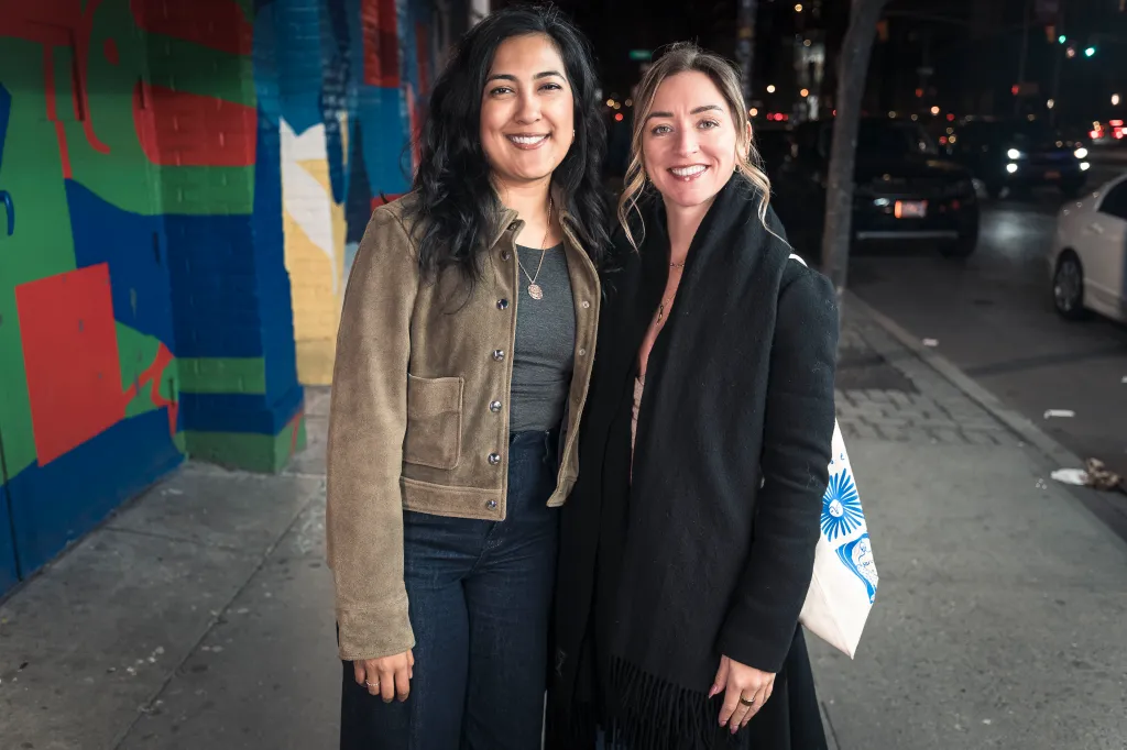 Two women smiling, Veronica with dark hair and Julie with blond hair, stand together on a city street.