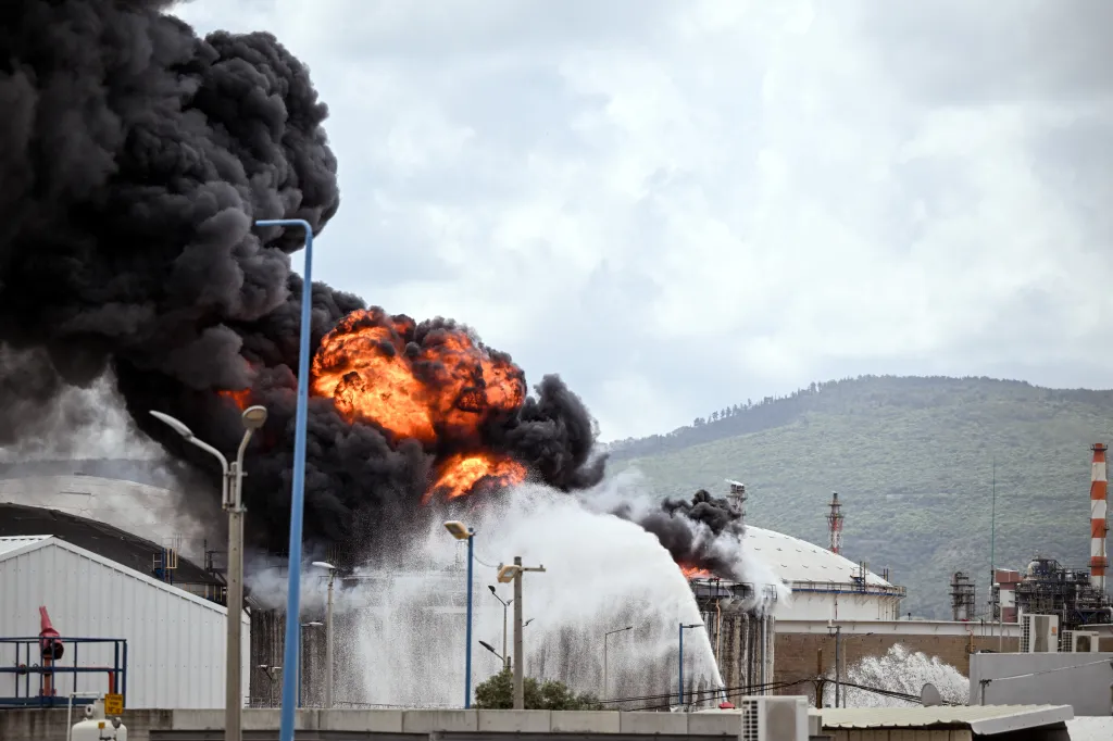 Large blaze with black smoke and water spraying on a fuel tanker at an oil refinery.