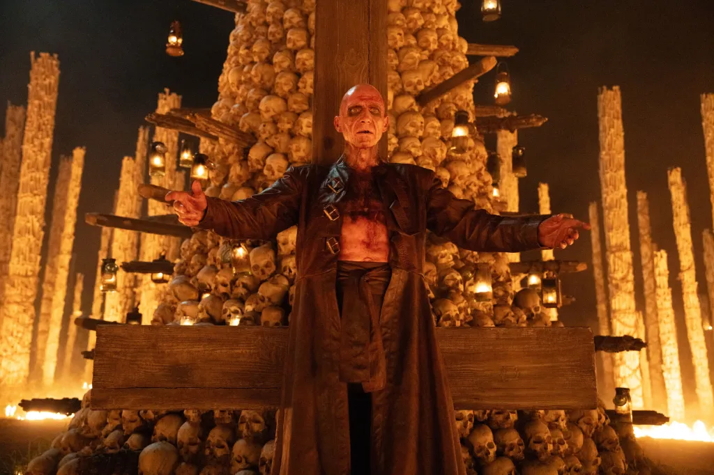 Ralph Fiennes as an older, bald, pale man with open arms, standing in front of a wall of skulls, with burning pillars and lanterns in the background.