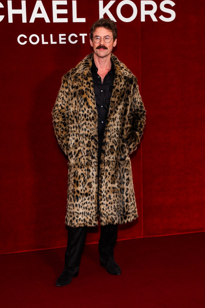 Brad Goreski wearing a leopard print coat over a black shirt and black pants, posing in front of a red Michael Kors sign.