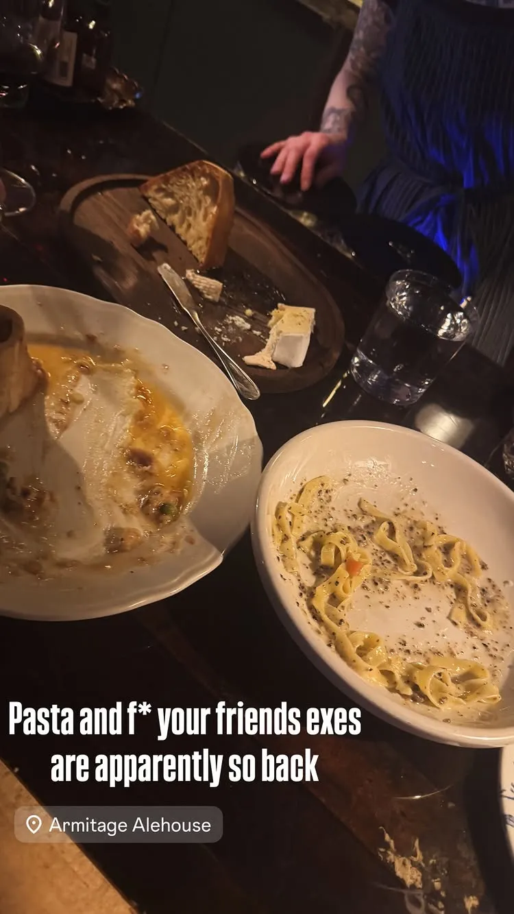 A messy table with leftover pasta, a cheese board with bread and cheese, and a glass of water, with a person's tattooed arm in the background.