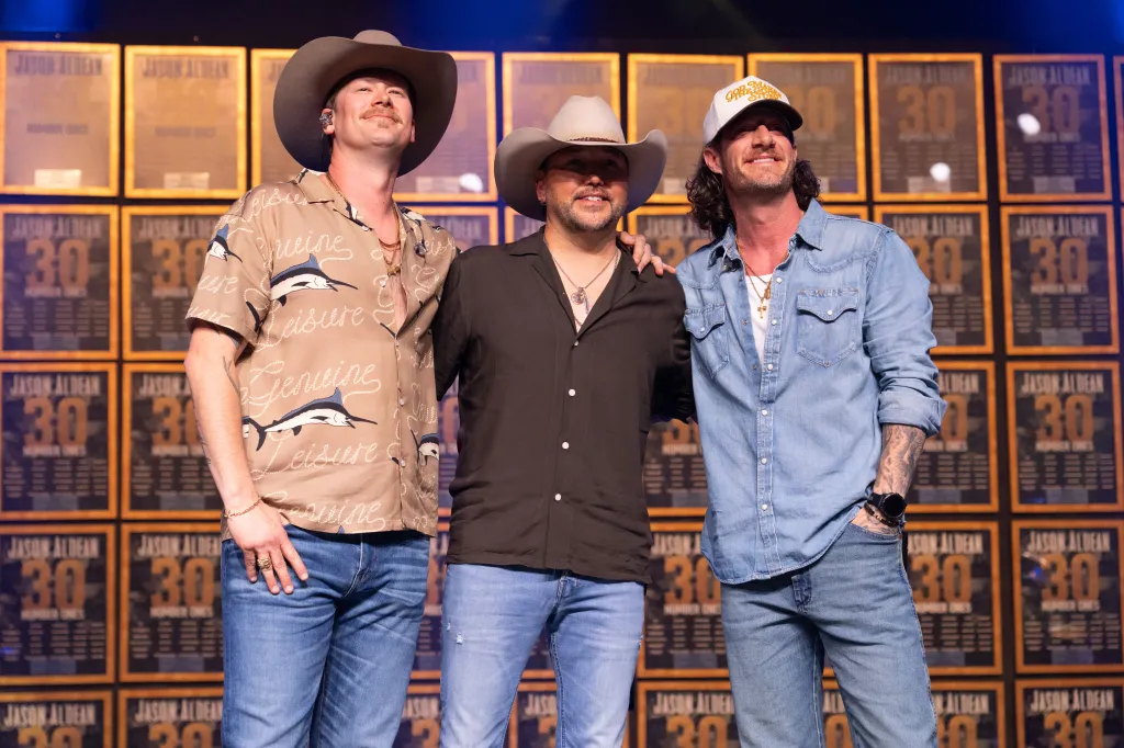 Brian Kelley, Jason Aldean, and Tyler Hubbard posing in front of plaques celebrating Jason Aldean's 31 number-one songs.