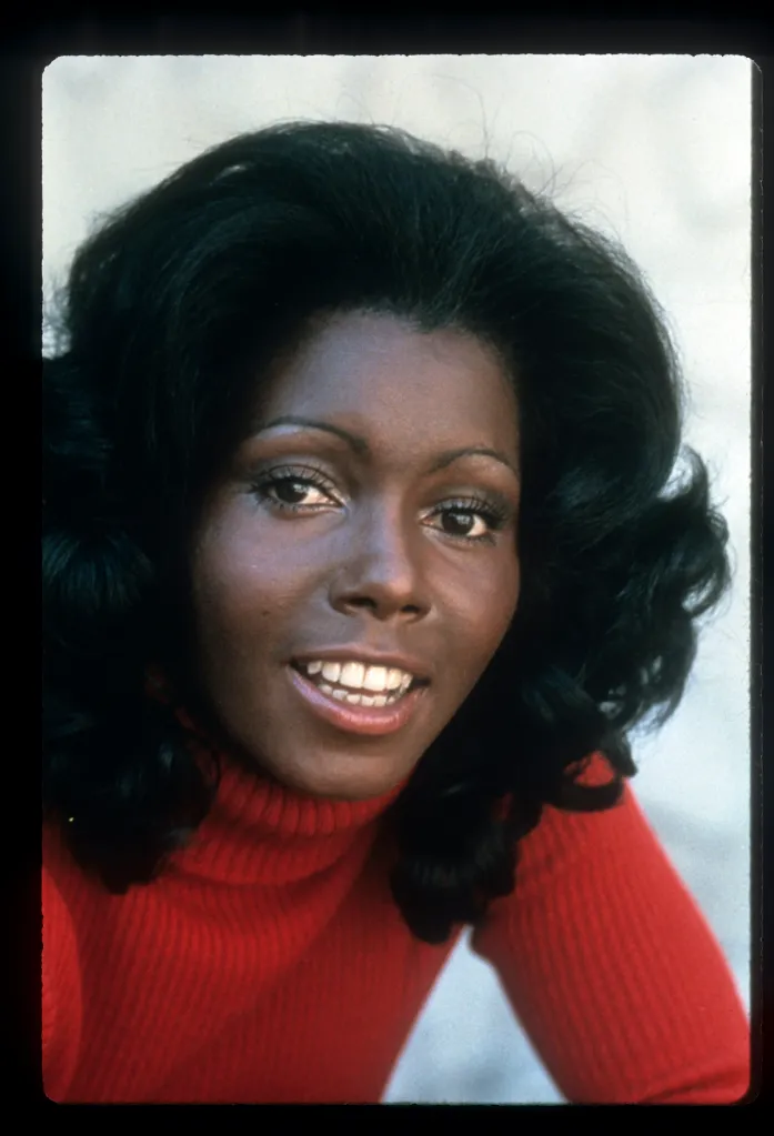 Judy Pace in a red turtleneck, smiling with a dark, voluminous hairstyle.