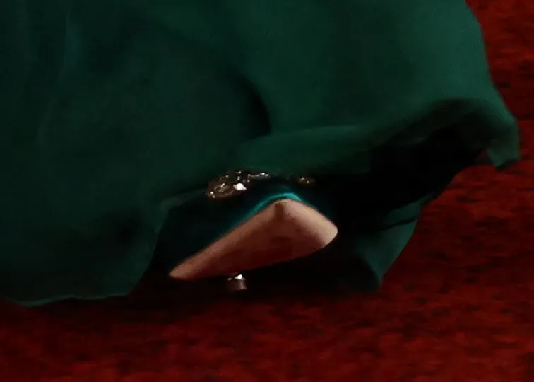 Catherine, Princess of Wales's beige high-heeled shoe beneath her dark green gown, with a diamond buckle on the shoe, on a red carpet.