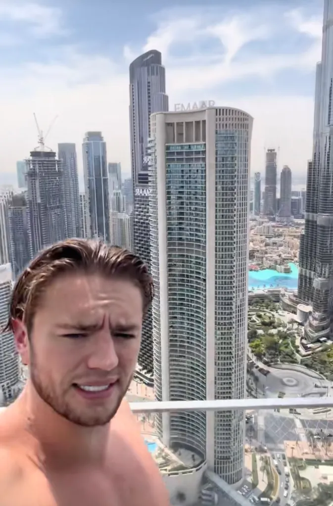 Mitchell Armstrong posing shirtless on a high-rise balcony in Dubai, with city buildings and a swimming pool visible in the background.