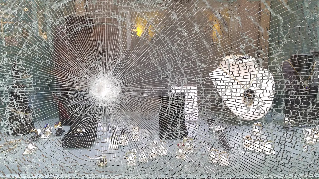 Shattered glass window of a jewelry store, with a display of watches, rings, and necklaces visible behind it.