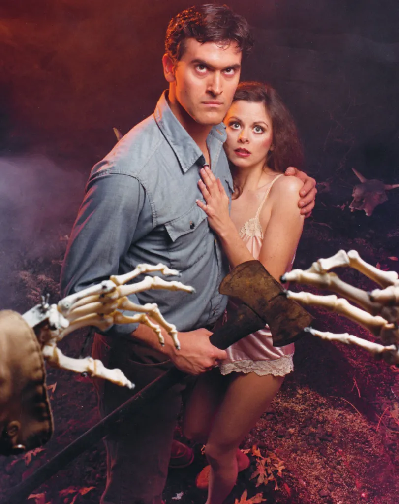Illustration of Bruce Campbell protecting Theresa Tilly, both look terrified, with a skeleton hand holding an axe in the foreground.