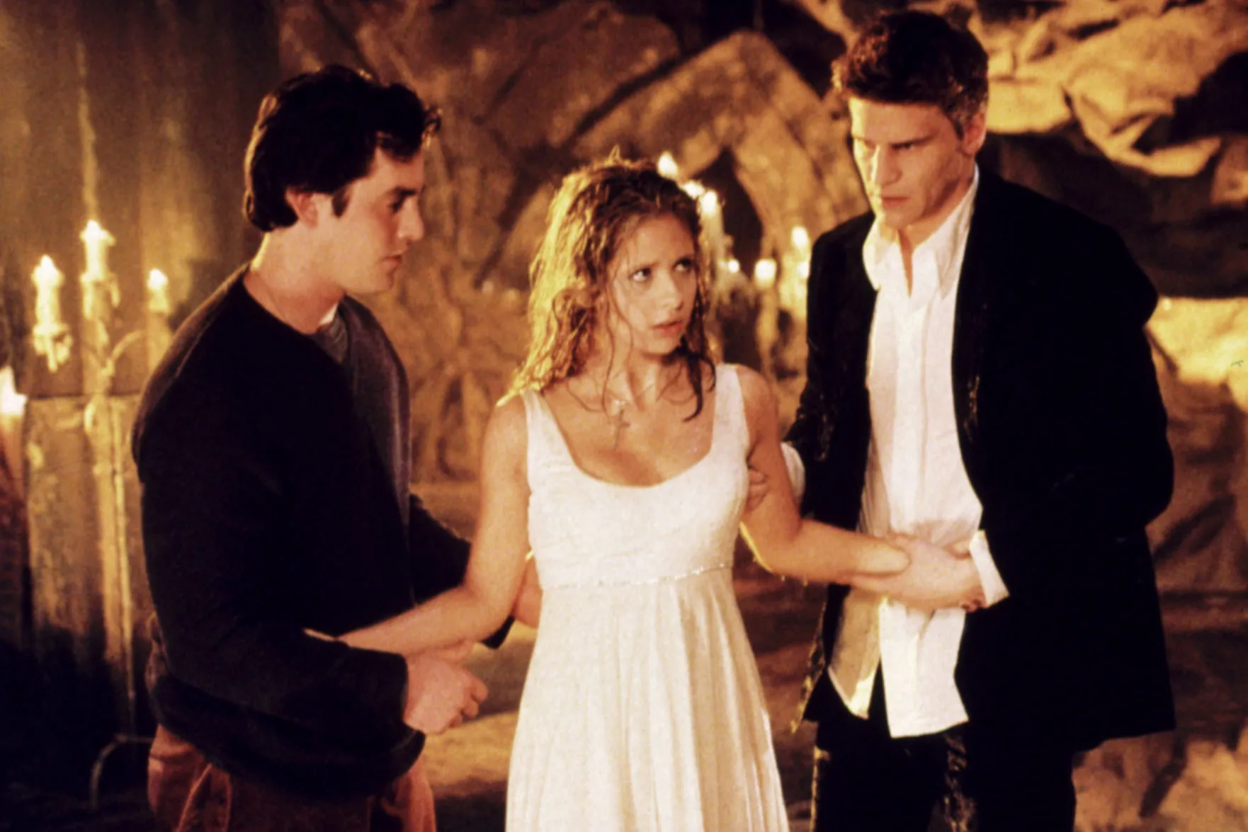 Sarah Michelle Gellar, caught between Nicholas Brendon and David Boreanaz, wearing a white dress, as they grab her arms.