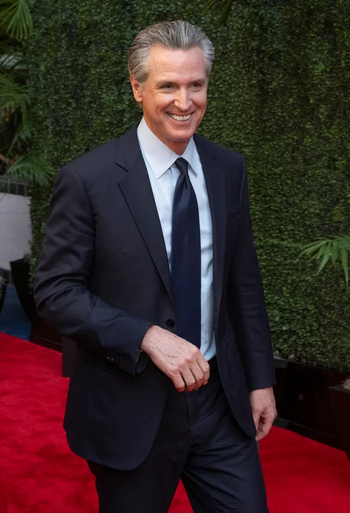 Gavin Newsom, California Governor, smiling in a navy suit with a lighter blue shirt and tie, walking on a red carpet.