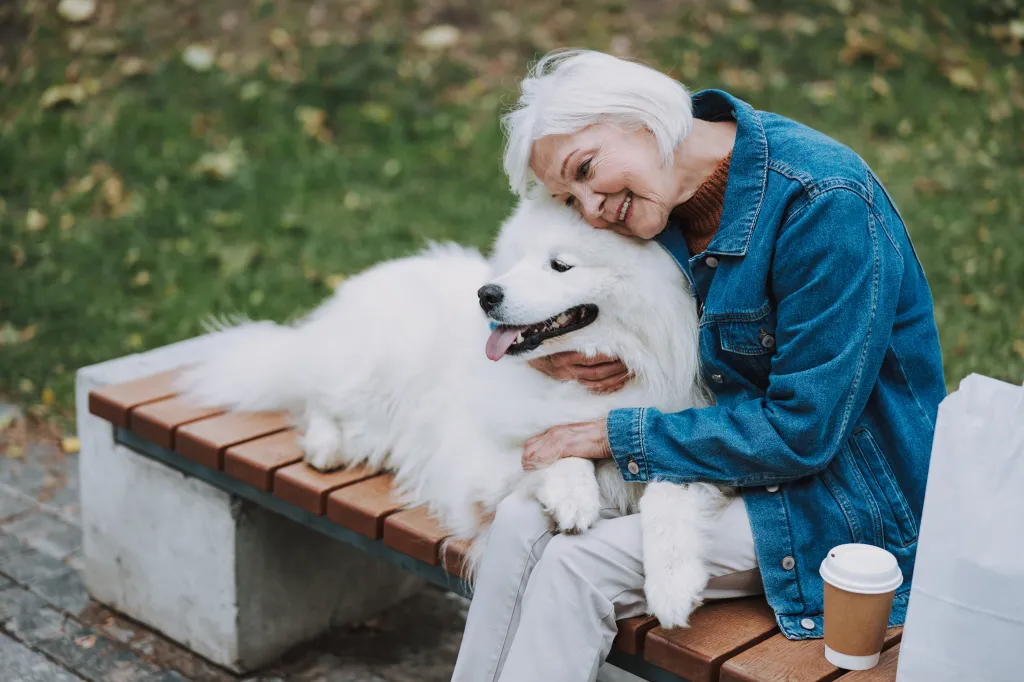 Happy woman in a denim jacket hugging her fluffy white dog on a park bench.