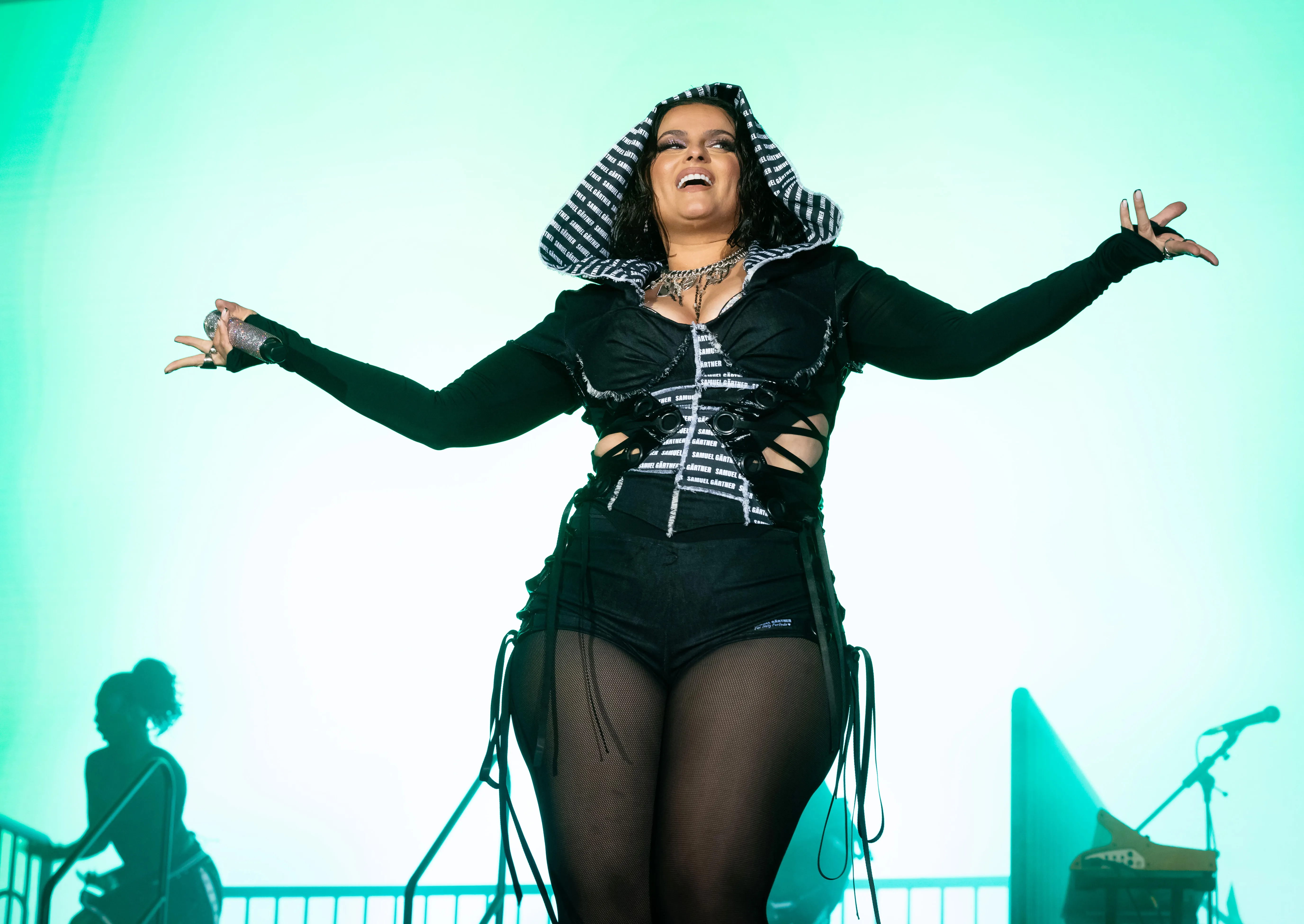 Nelly Furtado performing live on stage with her arms spread, wearing a dark hooded outfit and a large necklace.