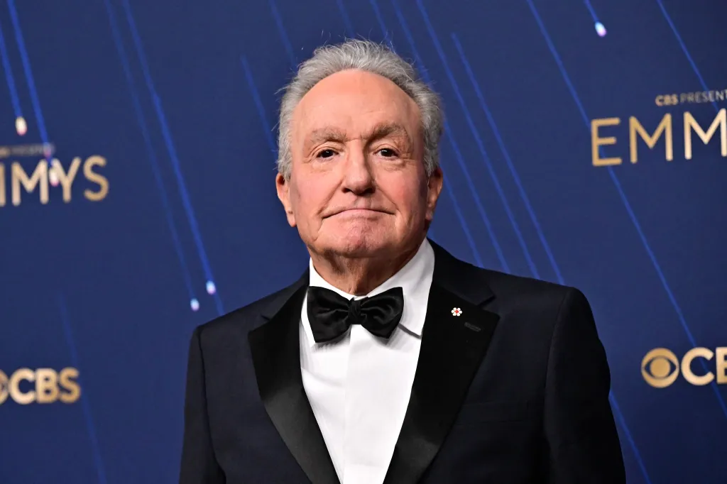 Lorne Michaels in a black tuxedo with a black bow tie and a flower pin on the lapel, standing in front of a blue background with the Emmys logo.