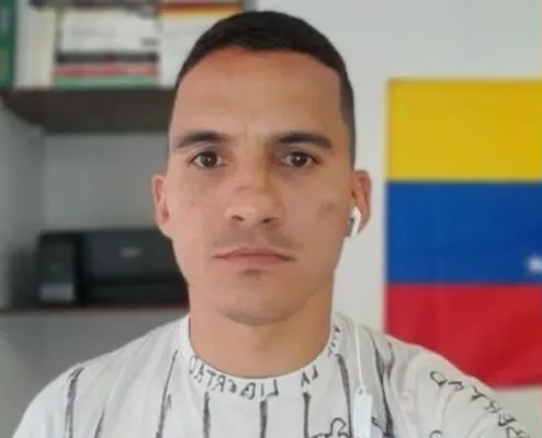 A man with short dark hair and a light-colored shirt, wearing white earbuds, looking directly at the camera with a Colombian flag in the background.