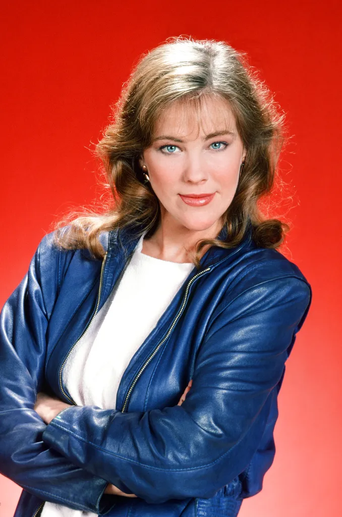 Catherine O'Hara in a blue leather jacket with her arms crossed, smiling at the camera.