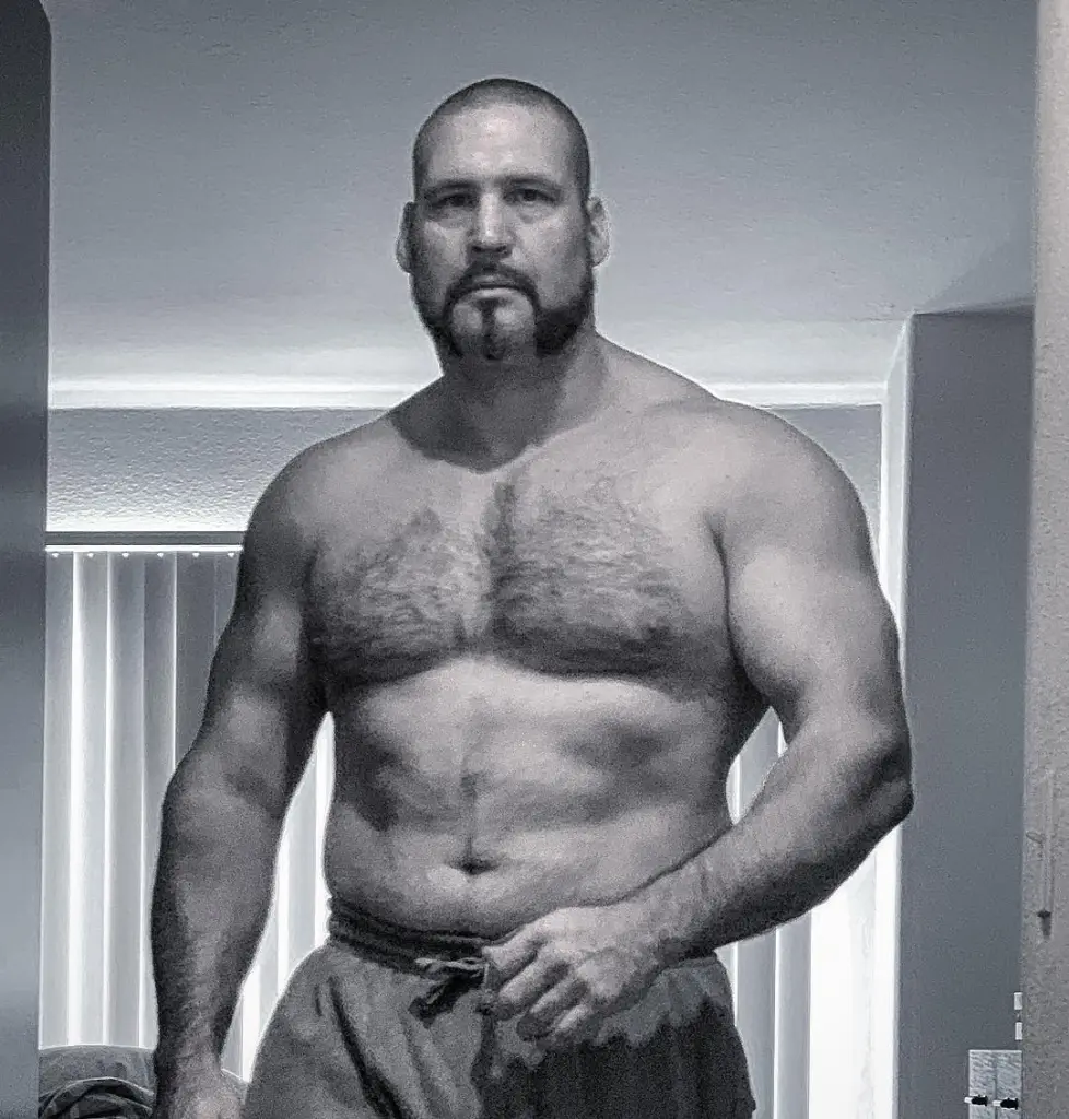 A shirtless, muscular man with a goatee looks directly at the camera.