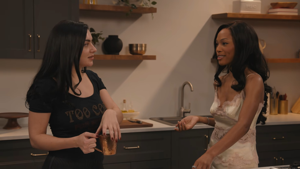 Charli XCX in a black shirt holding a copper mug, and another woman in a light-colored lace top, talking in a kitchen.