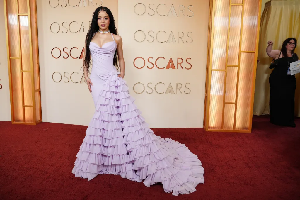 Chase Infiniti on the Oscars red carpet, wearing a lavender gown with a ruffled train.