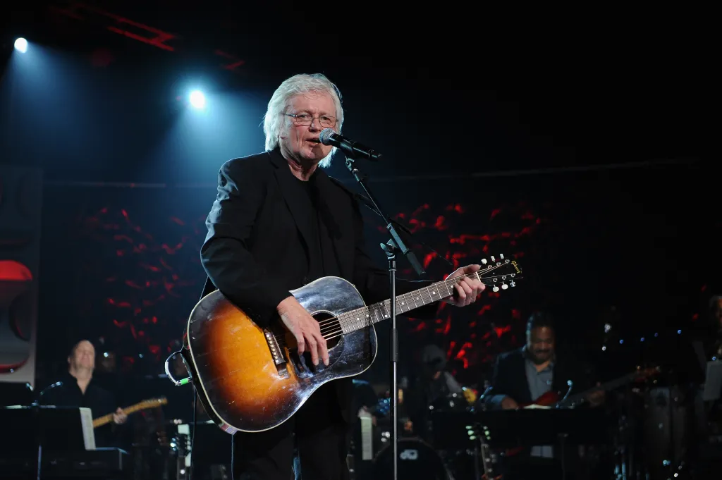 Chip Taylor performing on stage with an acoustic guitar.