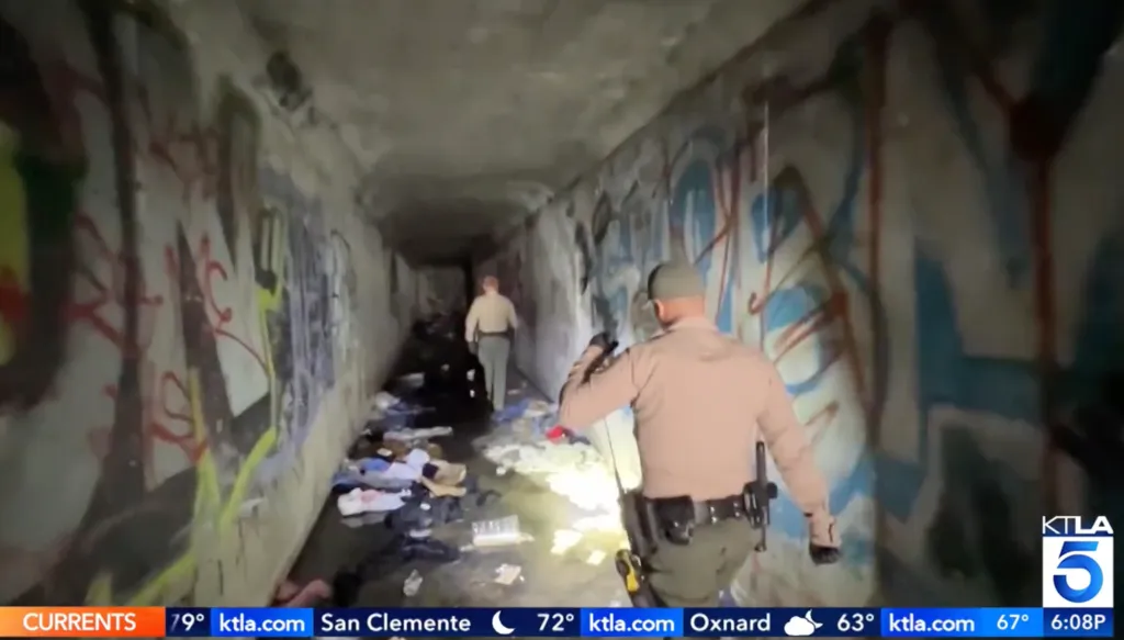 Two city officials inside a graffiti-covered storm drain with scattered belongings on the wet ground.