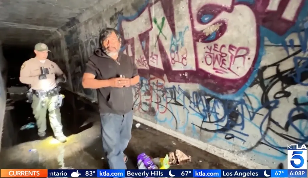 An officer and an unhoused man stand in a graffiti-covered storm drain with items on the ground.