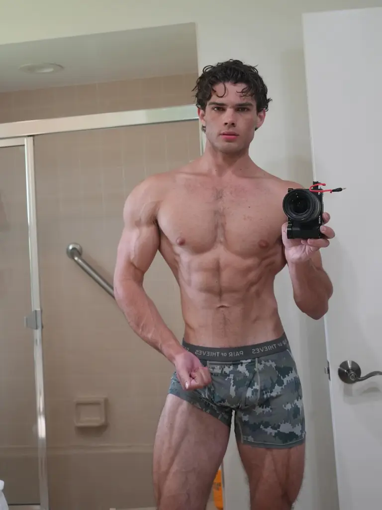 A man in camo boxer briefs holding a camera and flexing his biceps.