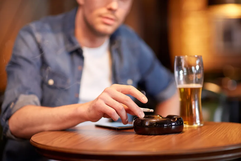 A man holding a lit cigarette over an ashtray, with a glass of beer on a table in front of him.